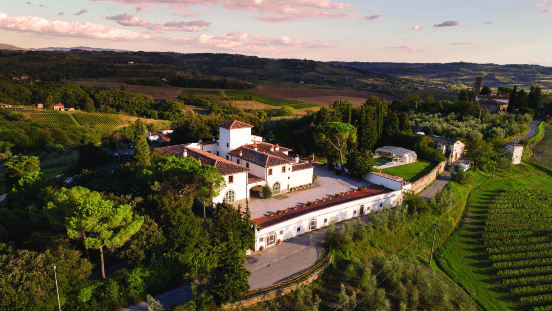 This image shows the Fattoria San Michele a Torri, which is a big winery. It is surrounded by vineyards. Taken from above, the image shows the winery in the middle, vineyards on all sides, and a skyline that is sunset-colored.
